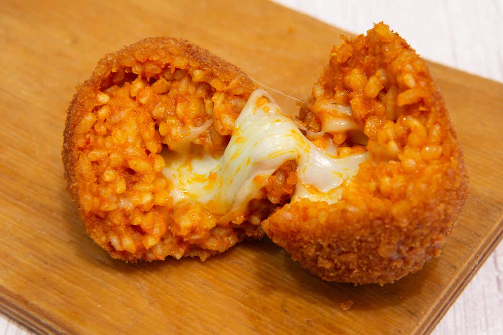 Un supplì spezzato a metà su un tagliere di legno, con la panatura dorata e croccante, il riso al pomodoro e la mozzarella filante al centro