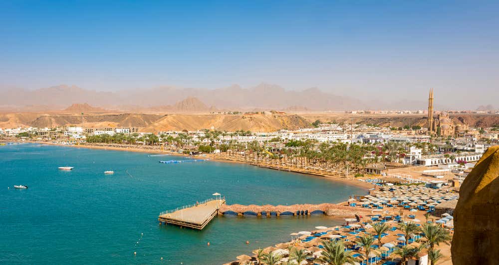 Vista panoramica della baia di Sharm el-Maya, a Sharm el-Sheikh, con l'acqua turchese e un pontile che si allunga nel mare. Sulla spiaggia ci sono file di ombrelloni e palme. Sullo sfondo si vedono gli edifici bianchi della città, il minareto della moschea Al Sahaba e le montagne del Sinai