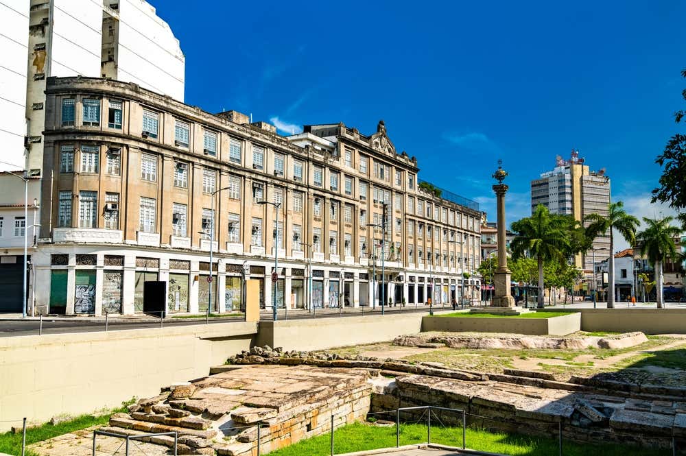 I resti in pietra del Cais do Valongo, l’antico molo nella zona portuale di Rio de Janeiro, conservati in un’area archeologica a cielo aperto. Sullo sfondo si vedono un grande palazzo storico color sabbia, una colonna commemorativa, palme ed edifici moderni sotto un cielo blu intenso