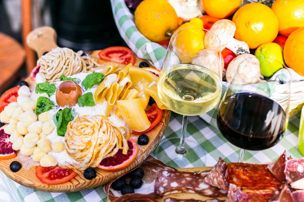 Una tavola apparecchiata all’esterno di un ristorante con una tovaglia a quadri verde e bianca, piena di cibo finto in mostra: pasta fresca, uova, mozzarelline, formaggio, basilico, pomodori e olive su un tagliere. Accanto ci sono due calici di vino, un tagliere di salumi e un cesto di agrumi e funghi