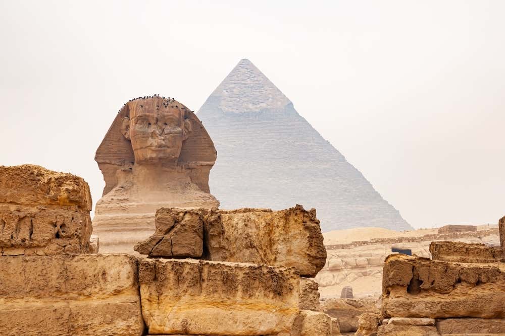 La Grande Sfinge di Giza, vista di fronte, con alcuni uccelli posati sulla testa. Alle sue spalle si alza la piramide di Chefren, che in cima conserva ancora un tratto del rivestimento originale. In primo piano ci sono blocchi di pietra calcarea erosi e il cielo è velato di foschia