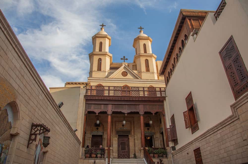 La facciata della Chiesa sospesa nel quartiere copto del Cairo, con due campanili gemelli sormontati da croci. In basso c'è un portico con colonne e un balcone in legno intagliato, e una scalinata sale verso l'ingresso. Ai lati della stretta corte si vedono pareti in pietra e finestre in legno lavorato a mashrabiya, sotto un cielo azzurro con nuvole bianche