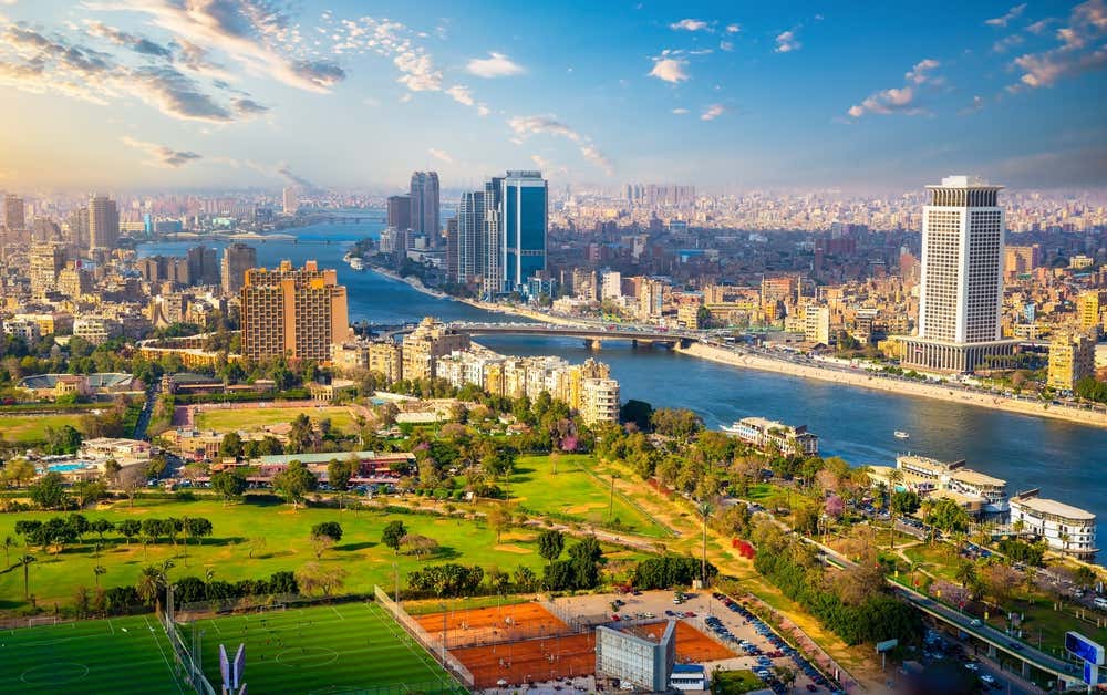 Vista panoramica del Cairo dall'alto, con il Nilo che attraversa la città tra grattacieli e palazzi. In primo piano ci sono i campi sportivi e il verde dell'isola di Gezira, mentre sullo sfondo si stende la città fino all'orizzonte, sotto un cielo azzurro con qualche nuvola