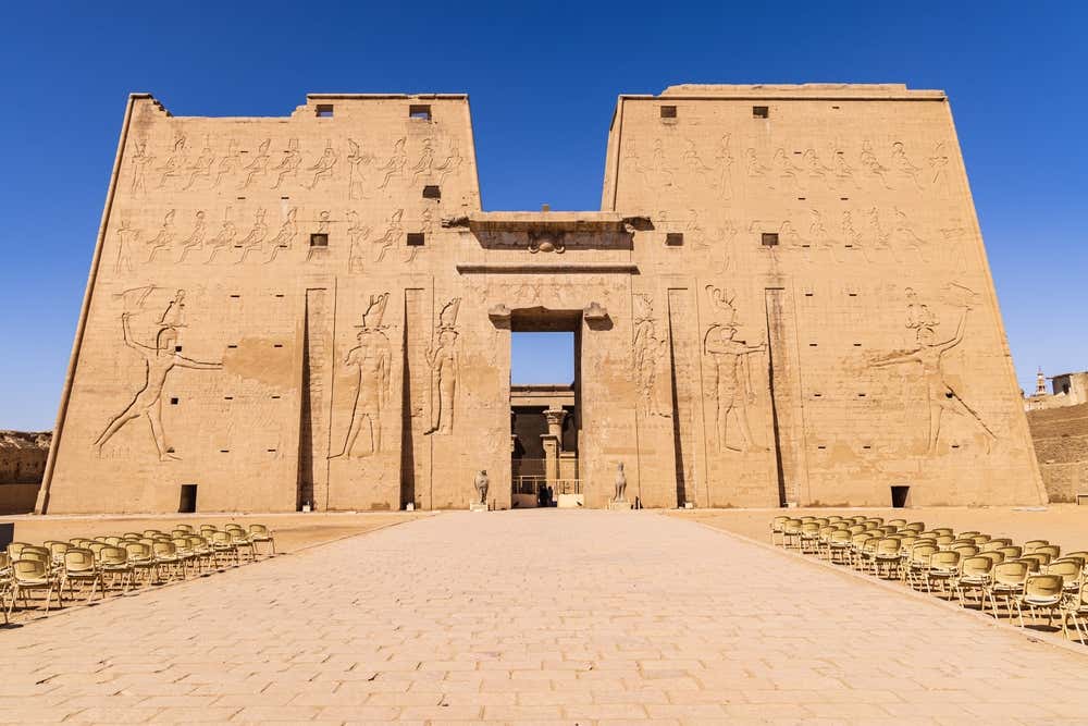 Il grande pilone d'ingresso del tempio di Edfu, dedicato al dio Horus. Le due torri in arenaria sono decorate con enormi bassorilievi del faraone che colpisce i nemici, sotto un cielo azzurro intenso. Al centro si apre il portale e ai lati del viale lastricato sono allineate file di sedie