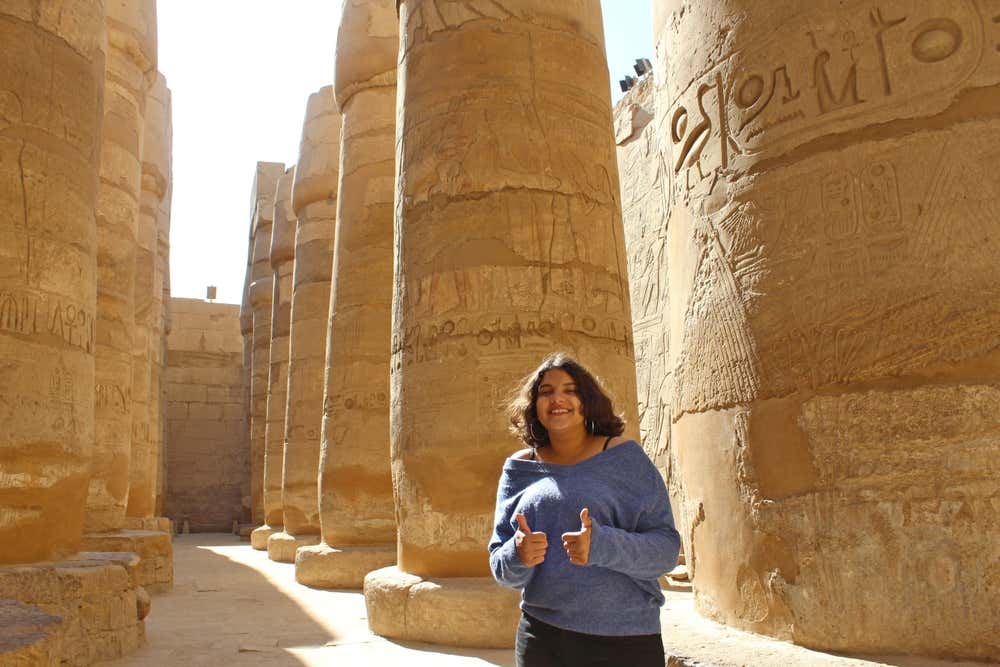 Una ragazza sorridente con un maglione blu fa il pollice in su tra le colonne della sala ipostila di Karnak, a Luxor. Le grandi colonne in pietra sono decorate con geroglifici e bassorilievi, e la luce del sole filtra tra di esse