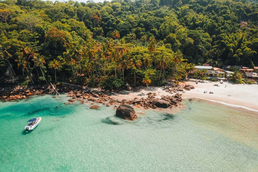 Vista aerea di una spiaggia di Ilha Grande, in Brasile, con l’acqua cristallina dalle sfumature turchesi e una barca a motore ancorata vicino alla riva. Sulla sabbia bianca ci sono grandi massi scuri e alcune case basse, mentre alle spalle della spiaggia sale una fitta foresta tropicale con palme