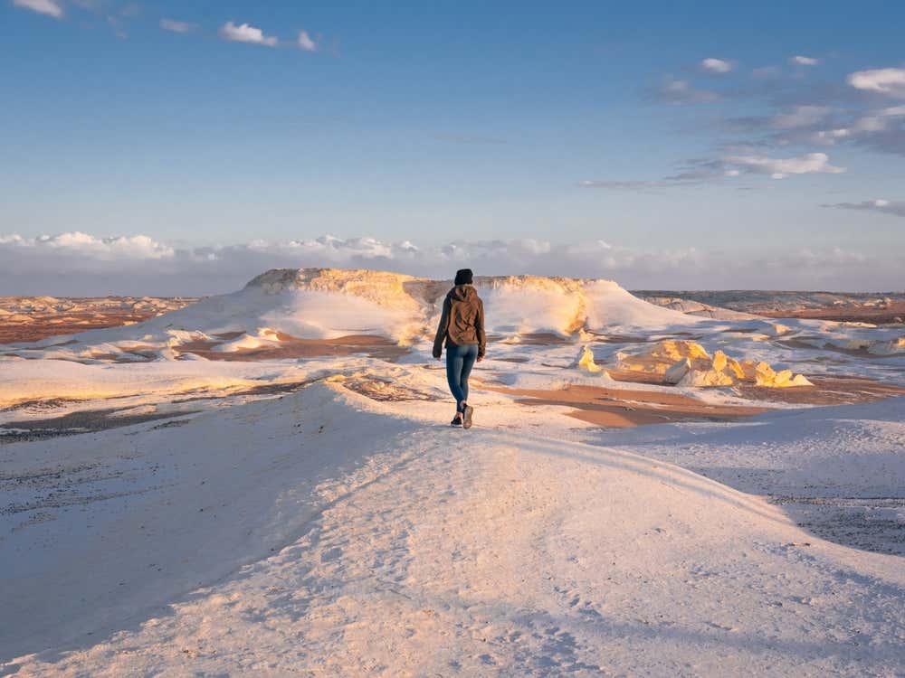 Una persona con giacca e jeans, vista di spalle, cammina su una cresta di roccia calcarea bianca nel Deserto Bianco, in Egitto. Intorno si stendono formazioni candide e sabbia color ocra, illuminate dalla luce calda del tramonto, sotto un cielo azzurro con qualche nuvola