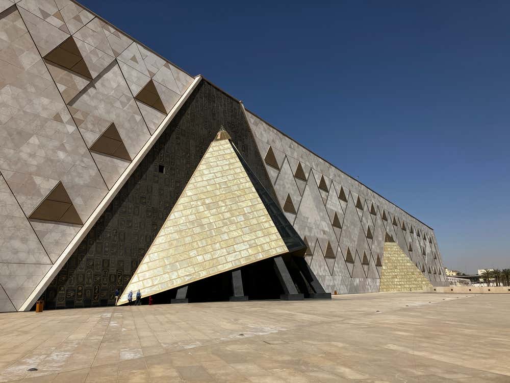 La facciata del Grande Museo Egizio, vicino a Giza: una lunga parete inclinata rivestita di pannelli triangolari in pietra chiara. Al centro c'è l'ingresso, coperto da una grande pensilina a forma di piramide con pannelli dorati, sotto un cielo blu intenso. Ai piedi dell'ingresso si vedono alcuni visitatori