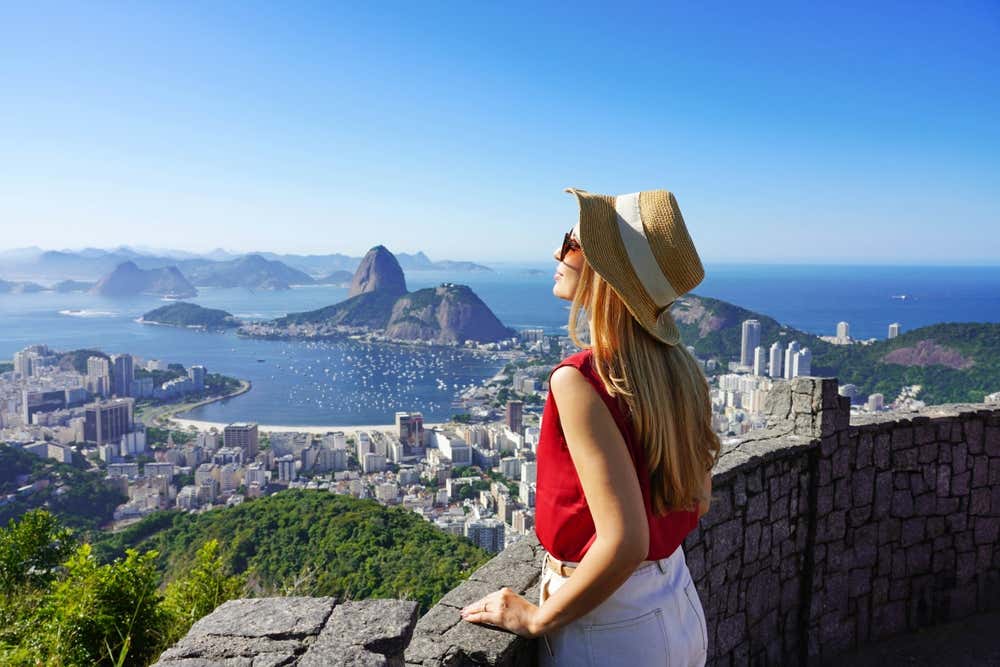 Una ragazza con un cappello di paglia e una camicetta rossa, appoggiata a un muretto in pietra, guarda il panorama di Rio de Janeiro. Sotto di lei si stendono la baia di Botafogo piena di barche, i palazzi della città e la spiaggia. Sullo sfondo si alza il Pan di Zucchero, circondato da isole e dal mare blu, sotto un cielo limpido
