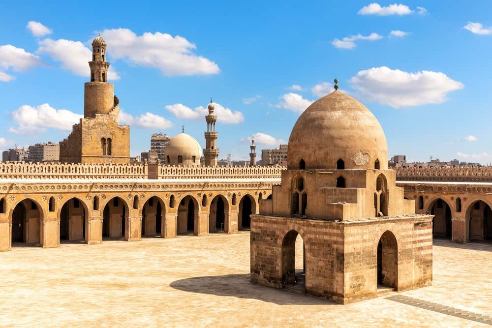 Il grande cortile della moschea di Ibn Tulun, al Cairo, circondato da portici con archi a sesto acuto e da una merlatura decorativa. In primo piano c'è la fontana per le abluzioni, coperta da una cupola. Sullo sfondo si vedono il minareto con la scala esterna a spirale, altri minareti e i palazzi della città, sotto un cielo azzurro con nuvole bianche