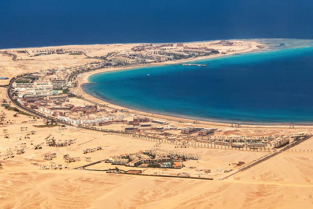 Vista aerea di una baia sul Mar Rosso, vicino a Hurghada. Il mare è turchese vicino alla riva e blu intenso al largo, e una lunga spiaggia di sabbia è circondata da resort e villaggi turistici. Tutto intorno si stende il deserto