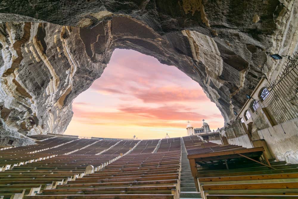 L'interno della chiesa rupestre di San Simone il conciatore, sul monte Mokattam al Cairo. File di panche in legno disposte ad anfiteatro scendono sotto un'enorme volta di roccia naturale, con venature chiare e scure. Dall'apertura della grotta si vede un cielo al tramonto sfumato di rosa e arancio, e sulla destra spuntano due piccole cupole con le croci