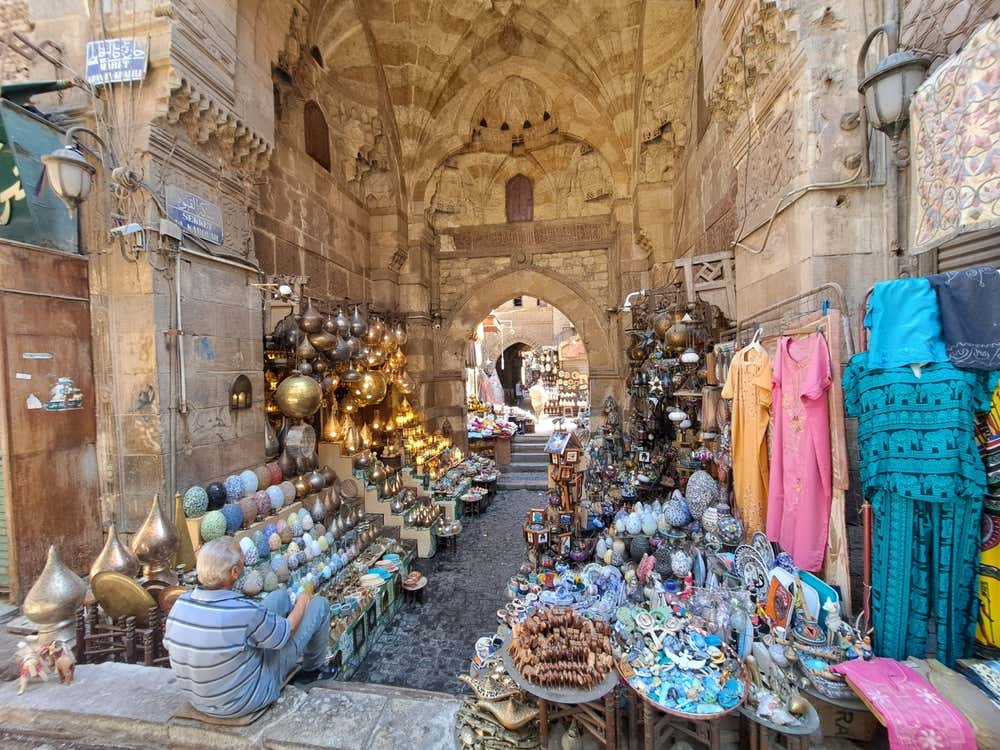 Una porta medievale in pietra ad arco nel bazar di Khan el-Khalili, al Cairo, con le bancarelle ai lati. Si vedono lampade in ottone, piatti in ceramica colorata, souvenir e abiti appesi. In primo piano un venditore è seduto accanto alla sua merce