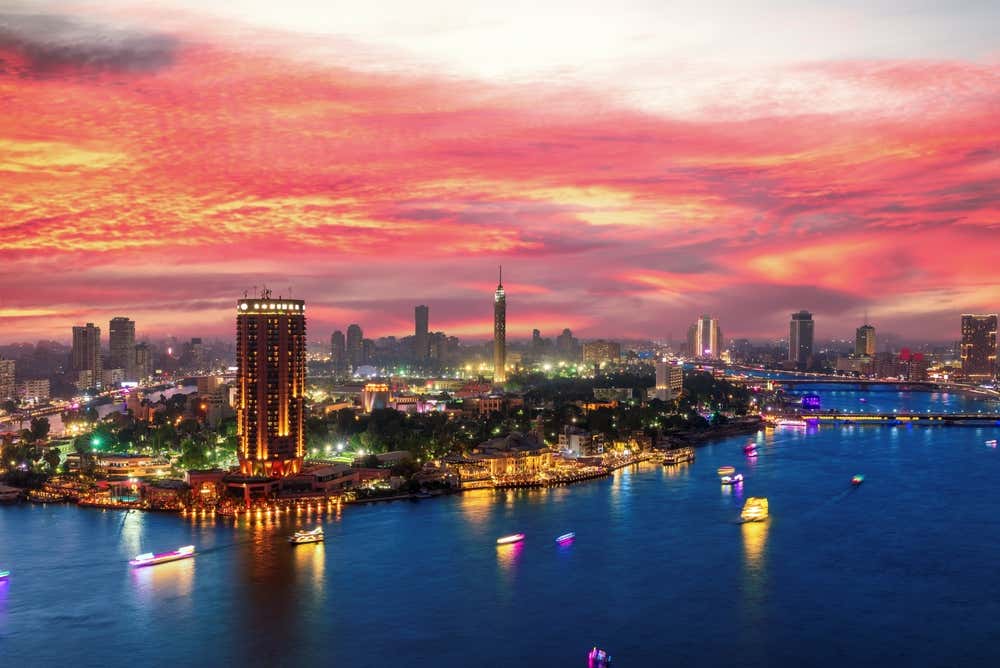 Il Cairo al tramonto, con il cielo tinto di rosa e arancio sopra lo skyline. Sul Nilo navigano battelli illuminati, mentre sulla riva si vedono l'isola di Gezira, un grattacielo illuminato e, al centro, la Torre del Cairo. Le luci della città si riflettono nell'acqua