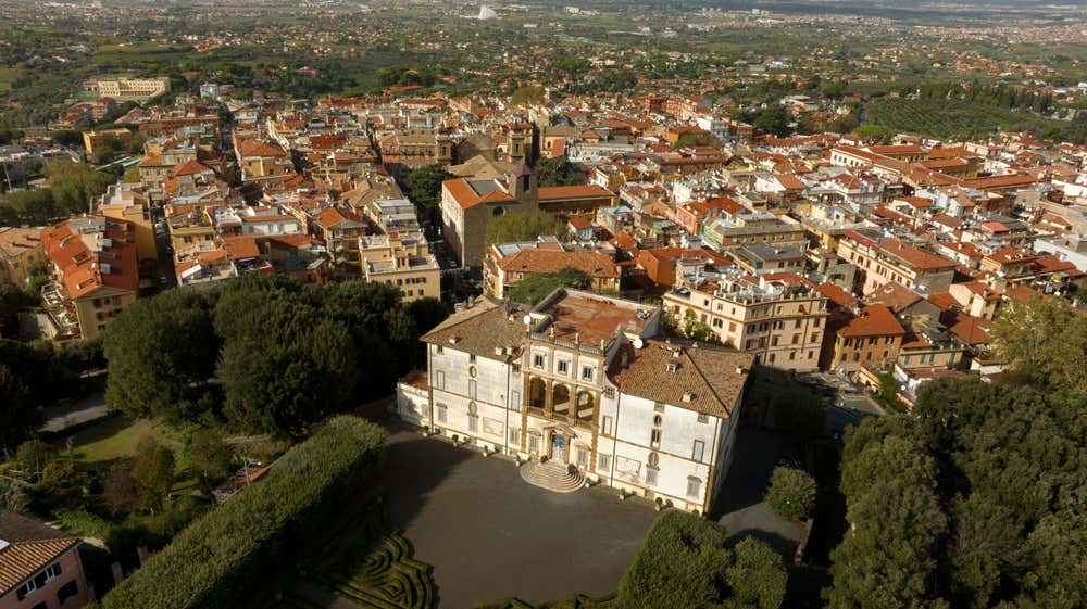 Vista aerea di Frascati, ai Castelli Romani: in primo piano una villa storica bianca con una loggia ad archi e una scalinata d’ingresso, circondata da giardini e siepi. Alle sue spalle si stendono i tetti rossi del paese e, in lontananza, la campagna verso Roma