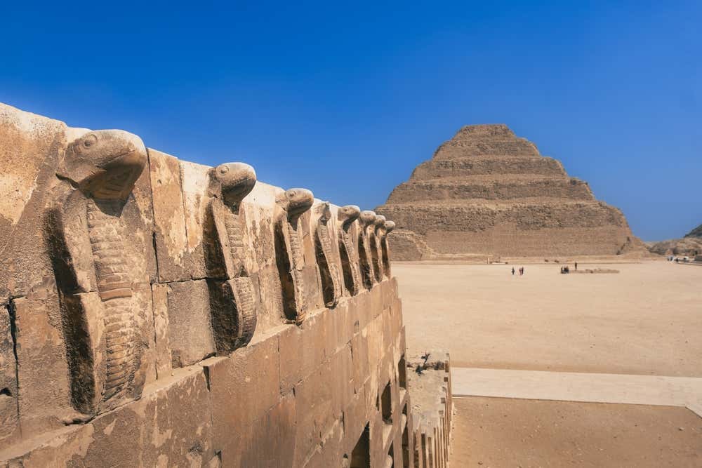 La piramide a gradoni di Djoser a Saqqara, in Egitto, vista sotto un cielo azzurro intenso. In primo piano c'è un muro in pietra calcarea decorato da un fregio di cobra scolpiti, mentre sul piazzale sabbioso davanti alla piramide si vedono alcuni visitatori