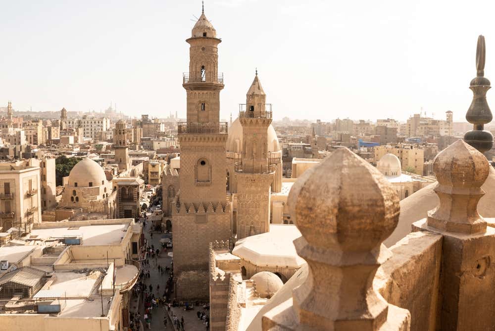 Vista dall'alto sul Cairo islamico, con i minareti in pietra e le cupole del centro storico. In basso a sinistra si vede una via stretta e affollata, mentre tutto intorno si stendono i tetti della città, avvolti da una leggera foschia. In primo piano ci sono le merlature decorative in pietra di un edificio