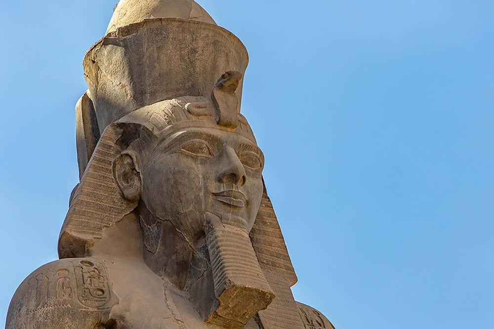 Il volto di una statua colossale di Ramses II scolpita nella pietra, vista dal basso contro un cielo azzurro. Il faraone ha la doppia corona dell'Alto e del Basso Egitto, il copricapo nemes, il cobra sulla fronte e la barba posticcia. Sulla spalla si vedono alcuni geroglifici incisi