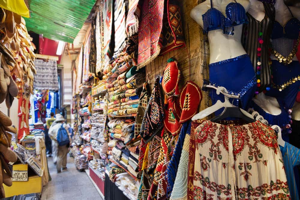 Un vicolo coperto del bazar di Khan el-Khalili, al Cairo, con le pareti piene di tessuti colorati, tappeti, borse ricamate e babbucce in pelle appese. Sulla destra alcuni manichini indossano costumi da danza del ventre blu con decorazioni dorate. In fondo al vicolo si vedono alcuni visitatori di passaggio