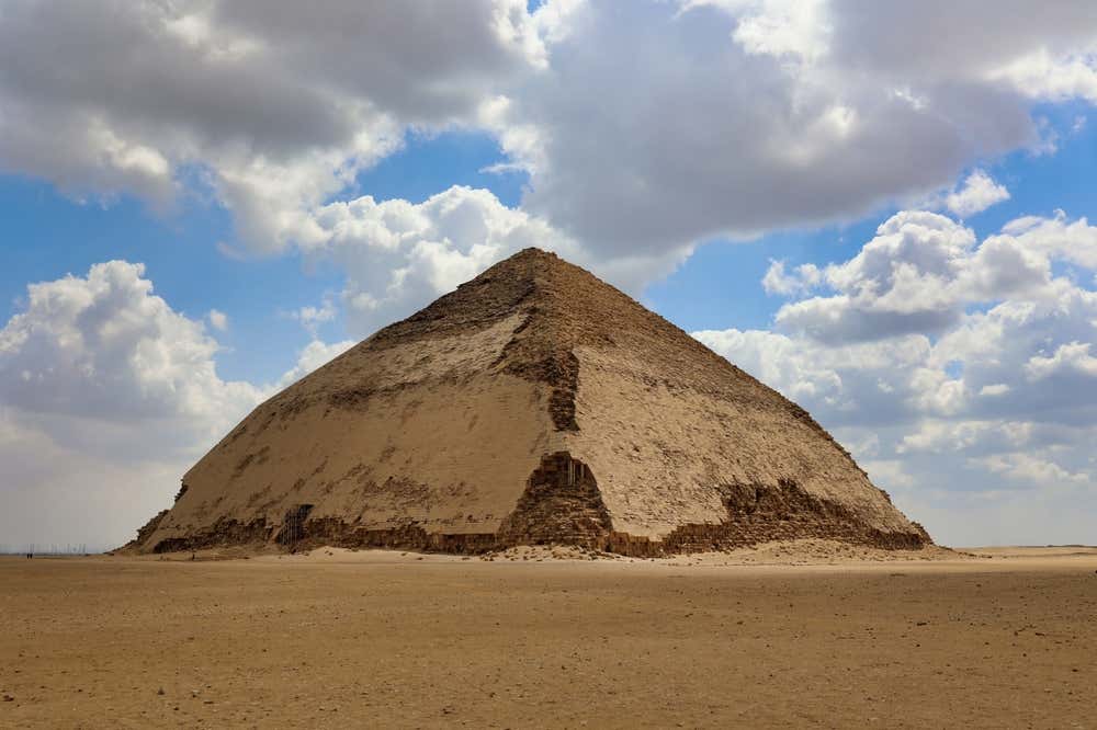 La piramide romboidale di Dahshur, in Egitto, sulla piana desertica. Gli spigoli cambiano inclinazione a metà altezza e su gran parte della superficie si vede ancora il rivestimento originale in pietra calcarea. Il cielo è azzurro, con grandi nuvole bianche