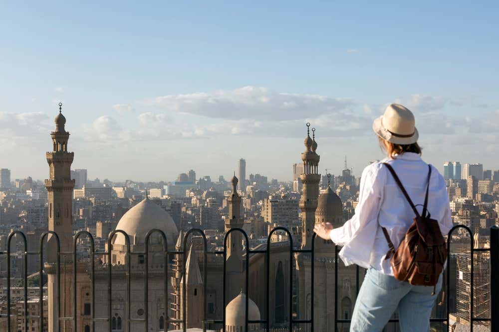 Una donna con un cappello di paglia, una camicia bianca e uno zaino in pelle, vista di spalle, guarda il Cairo da una terrazza con una ringhiera in ferro. Davanti a lei si alzano le cupole e i minareti delle moschee del sultano Hassan e di al-Rifai, e sullo sfondo la città si stende fino all'orizzonte, sotto un cielo azzurro