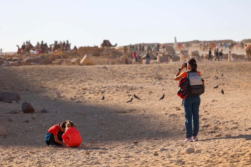 Una turista con uno zaino scatta una foto sulla piana di Giza, mentre due bambini con le magliette rosse giocano accovacciati sulla sabbia. Tutto intorno camminano alcuni uccelli, e sullo sfondo si vedono blocchi di pietra antichi, visitatori e una fila di cammelli, nella luce calda del pomeriggio