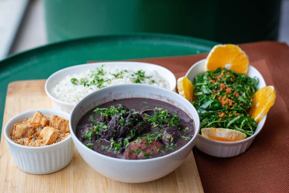 Una feijoada servita in una ciotola bianca, con fagioli neri, pezzi di carne di maiale e prezzemolo tritato. Intorno ci sono una ciotola di riso, una di cavolo tagliato fine con farofa e spicchi d’arancia, e una piccola ciotola di farofa con cubetti di carne fritta, su un tagliere di legno