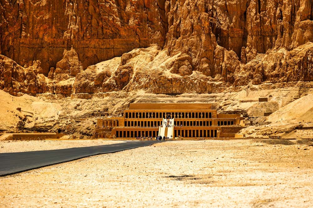 Il tempio di Hatshepsut a Deir el-Bahari, a Luxor, visto da lontano. Le sue terrazze a colonne, collegate da una rampa centrale, sono addossate a un'imponente parete di roccia dorata. In primo piano si stendono il piazzale sterrato e una strada asfaltata, mentre davanti al tempio si vedono alcuni visitatori