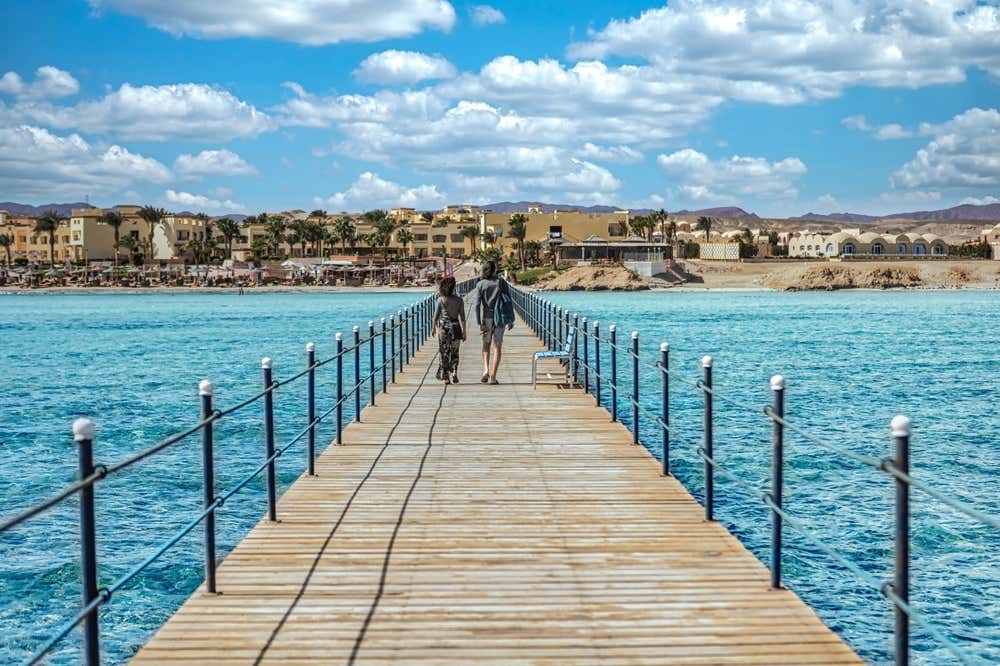 Due persone camminano su un lungo pontile in legno a Marsa Alam, sopra l'acqua turchese del Mar Rosso. Sullo sfondo si vedono una spiaggia con ombrelloni, i resort color sabbia tra le palme e le montagne del deserto, sotto un cielo azzurro con nuvole bianche