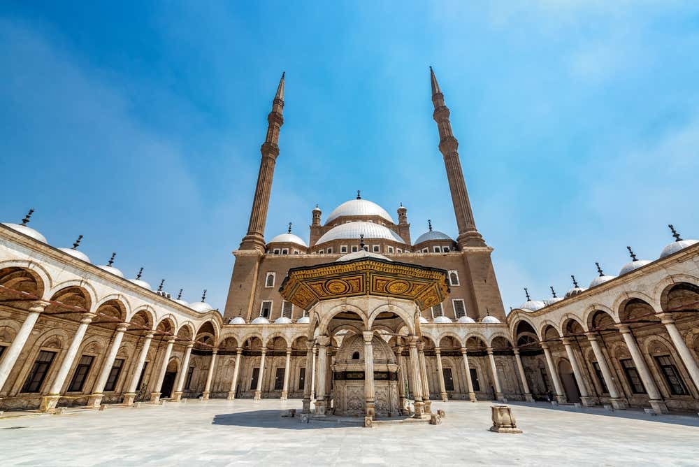 Il cortile della moschea di Muhammad Ali, nella cittadella di Saladino al Cairo. Al centro c'è la fontana per le abluzioni, con un tetto decorato in verde e oro. Tutto intorno corre un porticato ad arcate sormontato da piccole cupole, e sullo sfondo si alzano la cupola principale e i due minareti sottili, sotto un cielo azzurro