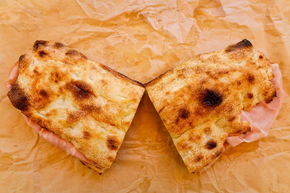 Due tranci di pizza bianca farciti con fette di mortadella, con la superficie dorata e qualche bruciatura del forno, appoggiati su un foglio di carta da forno spiegazzata
