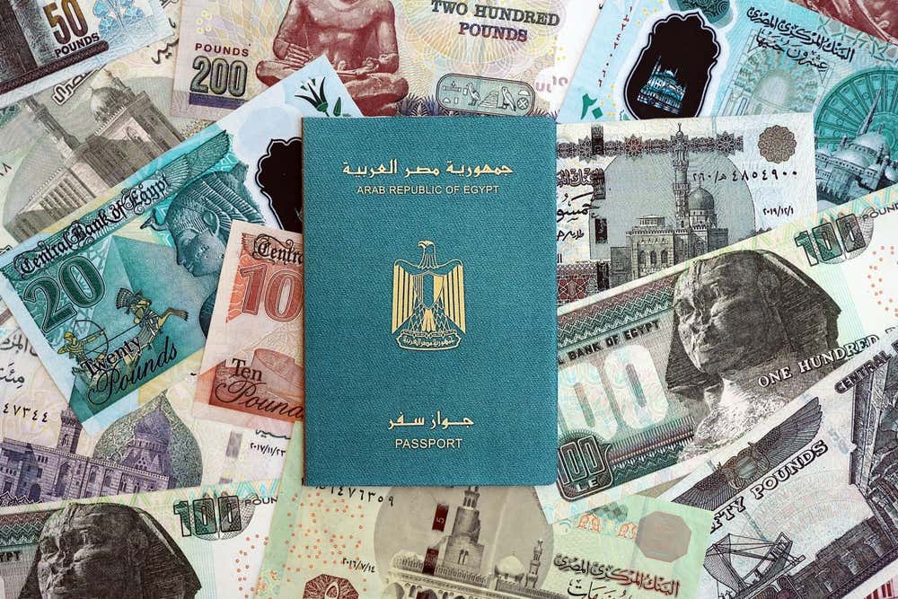 Un passaporto egiziano color verde acqua, con l'aquila dorata dello stemma, appoggiato su banconote egiziane di vari tagli. Sulle banconote si vedono la Sfinge, alcune moschee e figure dell'antico Egitto