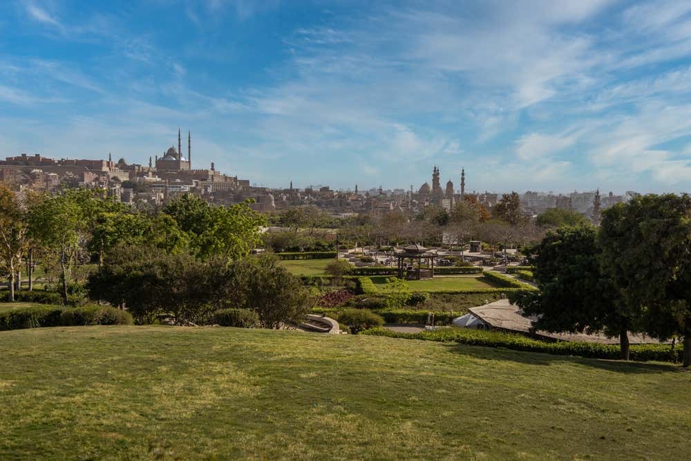 Il parco al-Azhar, al Cairo, con prati verdi, alberi e giardini geometrici con un padiglione al centro. Sullo sfondo si vede lo skyline della città, con la cittadella di Saladino e la moschea di Muhammad Ali a sinistra e altri minareti e cupole a destra, sotto un cielo azzurro con nuvole leggere