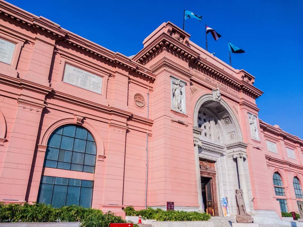 La facciata rosa del Museo Egizio in piazza Tahrir, al Cairo. L'ingresso principale è ad arco, decorato con statue in rilievo, e sopra di esso sventolano alcune bandiere. Davanti all'ingresso c'è una statua antica in pietra, sotto un cielo blu intenso