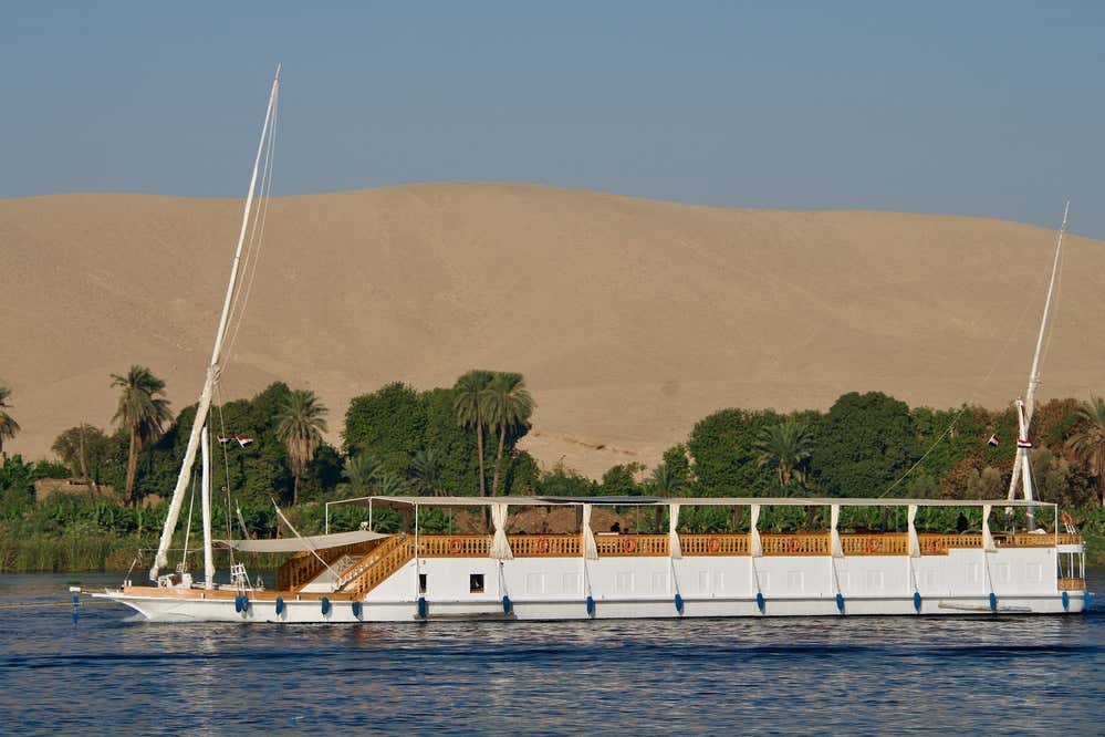 Una dahabiya, la tradizionale barca a vela del Nilo, naviga sul fiume. Lo scafo è bianco, con due alberi e un ponte coperto. Alle sue spalle si vedono una fascia di palme e vegetazione verde lungo la riva e, sullo sfondo, una grande duna di sabbia del deserto