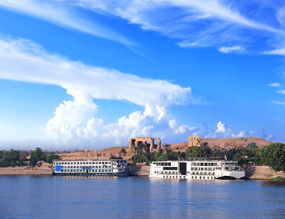 Due navi da crociera ormeggiate sulla riva del Nilo a Kom Ombo. Alle loro spalle, su una collinetta di sabbia, si vedono le colonne del tempio di Kom Ombo, circondate da palme e alberi. Il cielo è azzurro, con grandi nuvole bianche