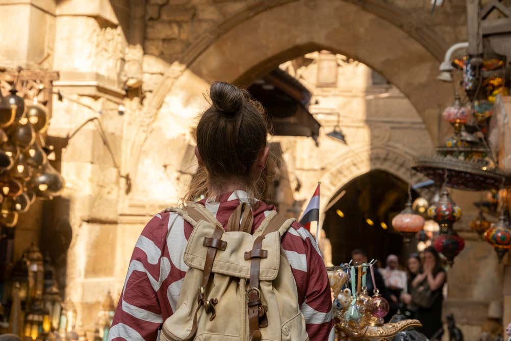 Una ragazza con uno zaino di tela beige, vista di spalle, cammina nel bazar di Khan el-Khalili al Cairo. Davanti a lei ci sono archi in pietra, lampade colorate appese e una piccola bandiera egiziana. Sullo sfondo si vedono altri visitatori