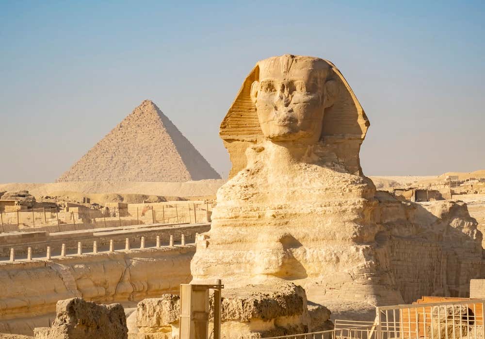 La Grande Sfinge di Giza, scolpita nella roccia calcarea, vista di tre quarti. Sullo sfondo si vede una delle grandi piramidi, sotto un cielo azzurro e terso.