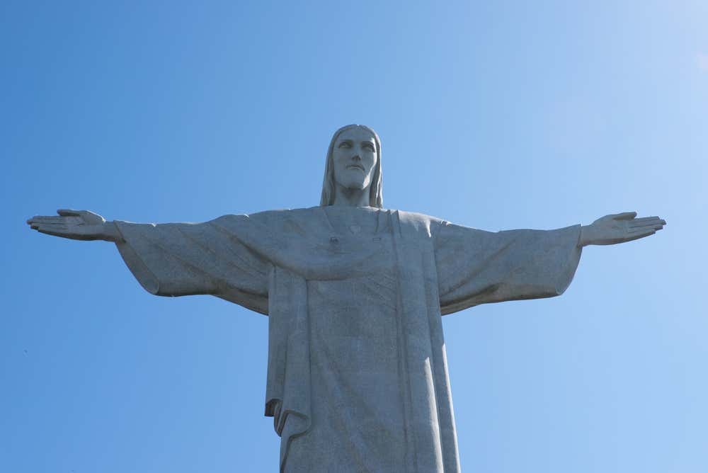 La statua del Cristo Redentore sul Corcovado, a Rio de Janeiro, vista frontalmente dal basso. La figura in pietra chiara ha le braccia aperte e indossa una lunga tunica, contro un cielo azzurro limpido