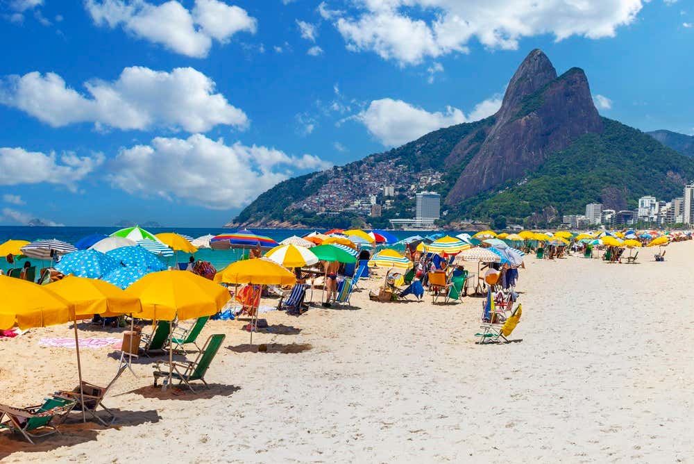 La spiaggia di Ipanema, a Rio de Janeiro, piena di ombrelloni colorati e sedie a sdraio sulla sabbia chiara. Sullo sfondo si alzano i due picchi del Morro Dois Irmãos e, sul fianco della collina, le case della favela di Vidigal, sotto un cielo azzurro con nuvole bianche