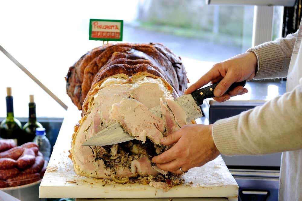 Un norcino affetta con un grosso coltello una porchetta intera su un tagliere bianco: la crosta è dorata e croccante, la carne rosata e condita con erbe aromatiche. Accanto si vedono un cartellino con la scritta “Porchetta”, alcune salsicce e bottiglie di vino