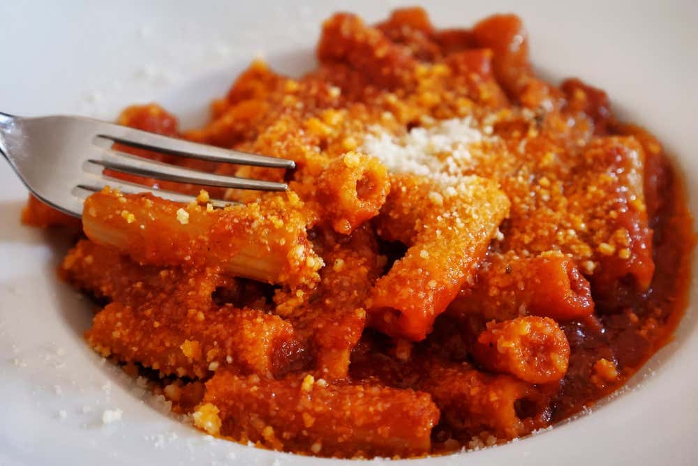 Un piatto di rigatoni all’amatriciana con sugo di pomodoro, pezzetti di guanciale e una generosa spolverata di pecorino grattugiato, in un piatto fondo bianco. Sulla sinistra una forchetta sta prendendo la pasta