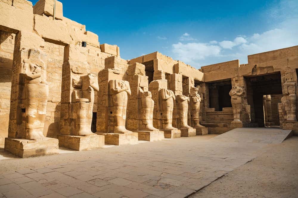 Il cortile di un tempio nel complesso di Karnak, a Luxor, delimitato da una fila di pilastri in arenaria. Su ogni pilastro è scolpita una grande statua del faraone in forma di Osiride, con le braccia incrociate sul petto. Sopra il cortile c'è un cielo azzurro con qualche nuvola