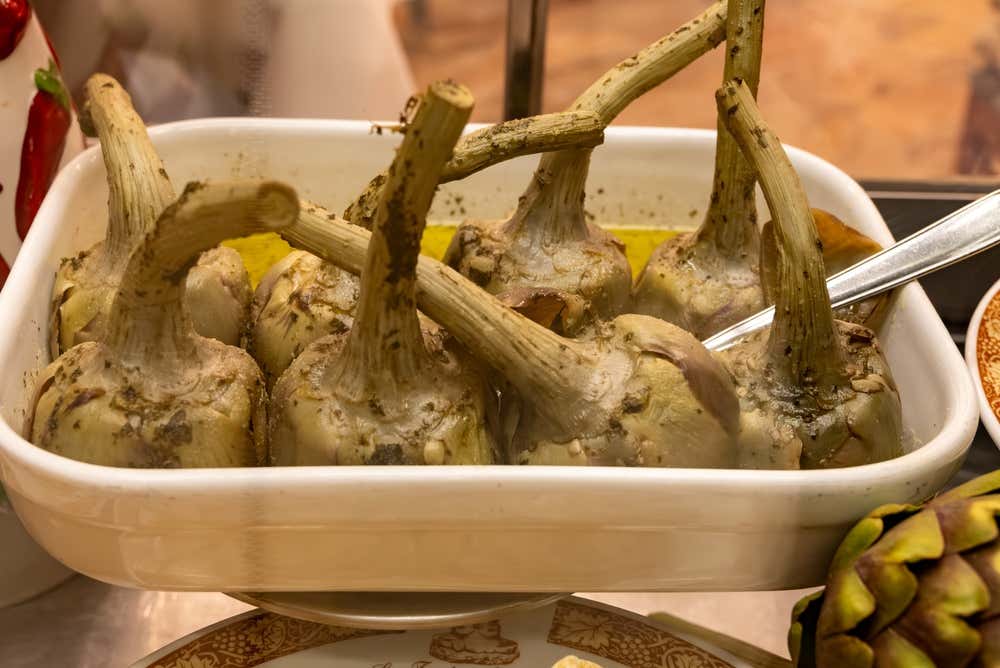 Una pirofila bianca piena di carciofi alla romana, cotti interi a testa in giù con i gambi rivolti verso l’alto, nell’olio con erbe aromatiche tritate. Accanto si vede un carciofo crudo e sullo sfondo alcuni peperoncini rossi