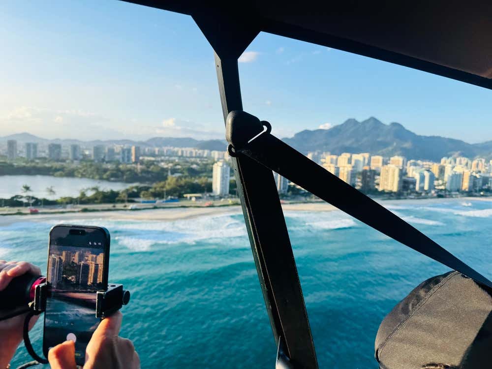 Una persona fotografa con lo smartphone il panorama di Rio de Janeiro da un elicottero senza porte. Sotto si vedono il mare turchese con le onde che si infrangono sulla spiaggia, una laguna, palazzi moderni e montagne sullo sfondo, nella luce calda del pomeriggio
