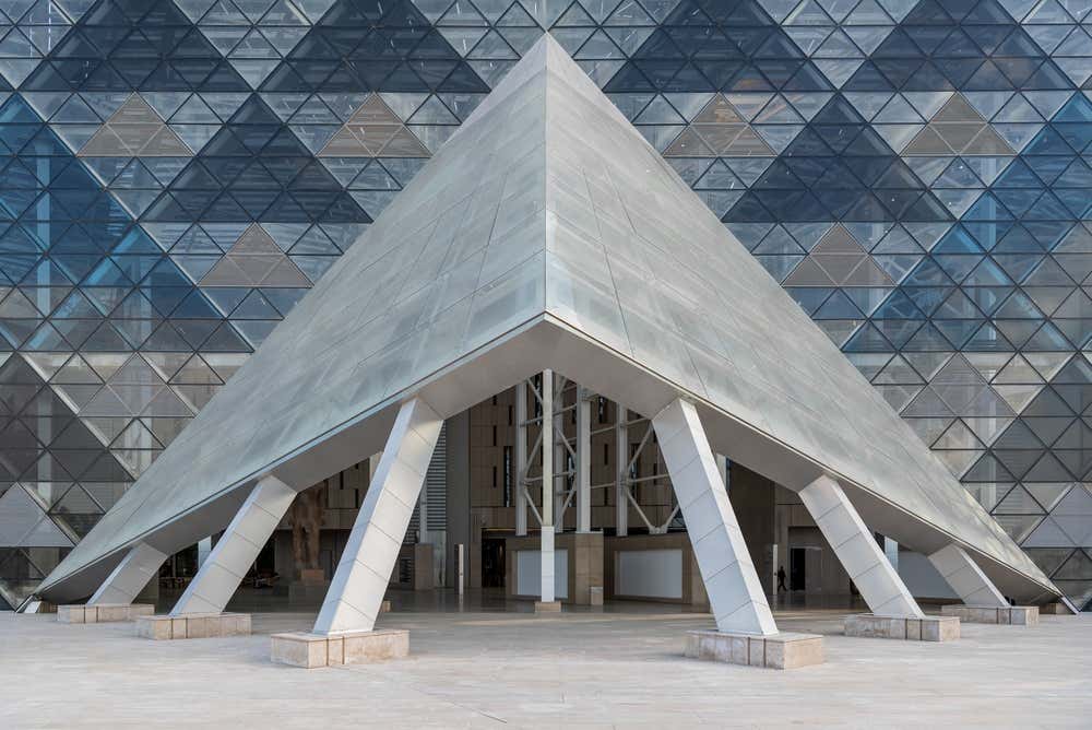 L'ingresso del Grande Museo Egizio, vicino alle piramidi di Giza: una grande pensilina a forma di piramide poggia su pilastri inclinati. Alle sue spalle c'è una facciata in vetro composta da pannelli triangolari