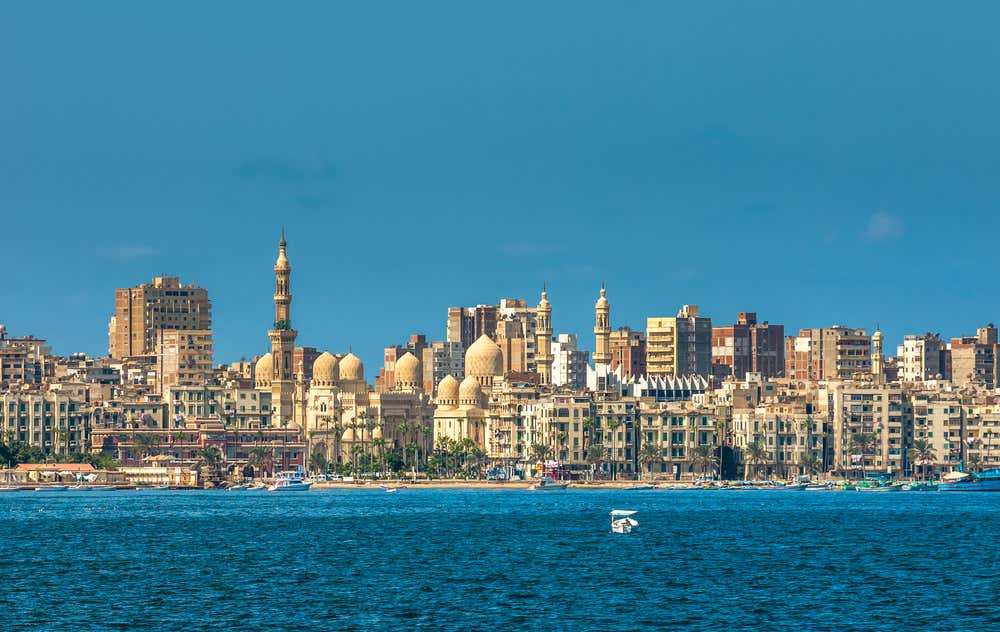 Il lungomare di Alessandria d'Egitto visto dal mare. In primo piano c'è l'acqua blu intenso e sullo sfondo si allinea una fila di palazzi. Tra gli edifici spicca la moschea di Abu al-Abbas al-Mursi, con il suo alto minareto e le cupole color sabbia