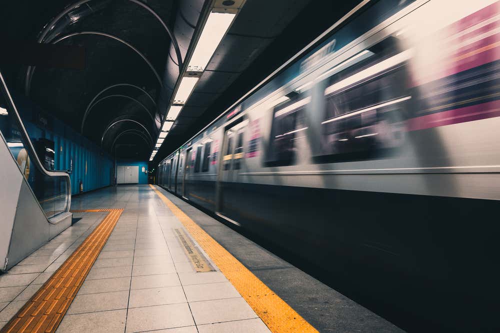 La banchina di una stazione della metropolitana a Copacabana, a Rio de Janeiro, con un treno argentato in movimento ripreso con l’effetto mosso. Il pavimento è chiaro, con le strisce gialle di sicurezza e i percorsi tattili, e sulla sinistra si vede una scala mobile accanto a una parete blu