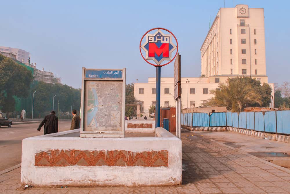 L'ingresso della stazione Sadat della metropolitana del Cairo, con il cartello rosso e blu della metro e la scritta in arabo. Accanto alla scala d'accesso c'è un pannello con la mappa della rete e il nome della stazione. Sullo sfondo si vedono un grande edificio chiaro con un orologio, alcuni passanti e una strada, in una giornata velata di foschia