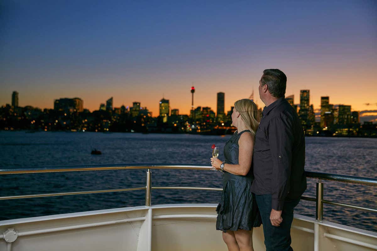 Horizonte de Sydney ao pôr do sol com um casal na beira de um barco admirando a paisagem.
